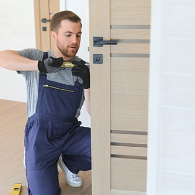 Installation of a lock on the front wooden entrance door. Portrait of young locksmith workman in blue uniform installing door knob. Professional repair service. Maintenance Concept Installation of a lock on the front wooden entrance door. Portrait of young locksmith workman in blue uniform installing door knob. Professional repair service. Maintenance Concept.